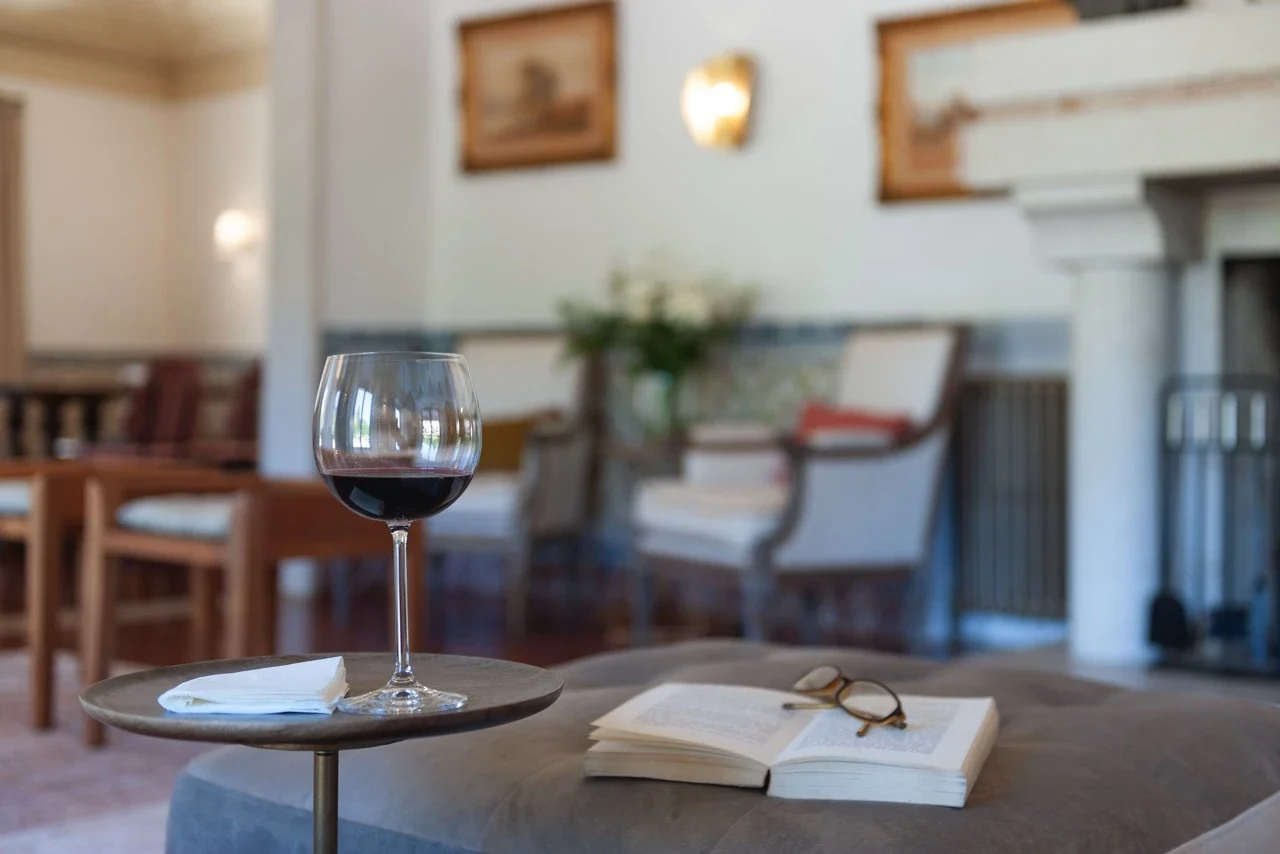 A photo of a glass of wine and an open book with glasses resting on it in the main room of the Manor house.